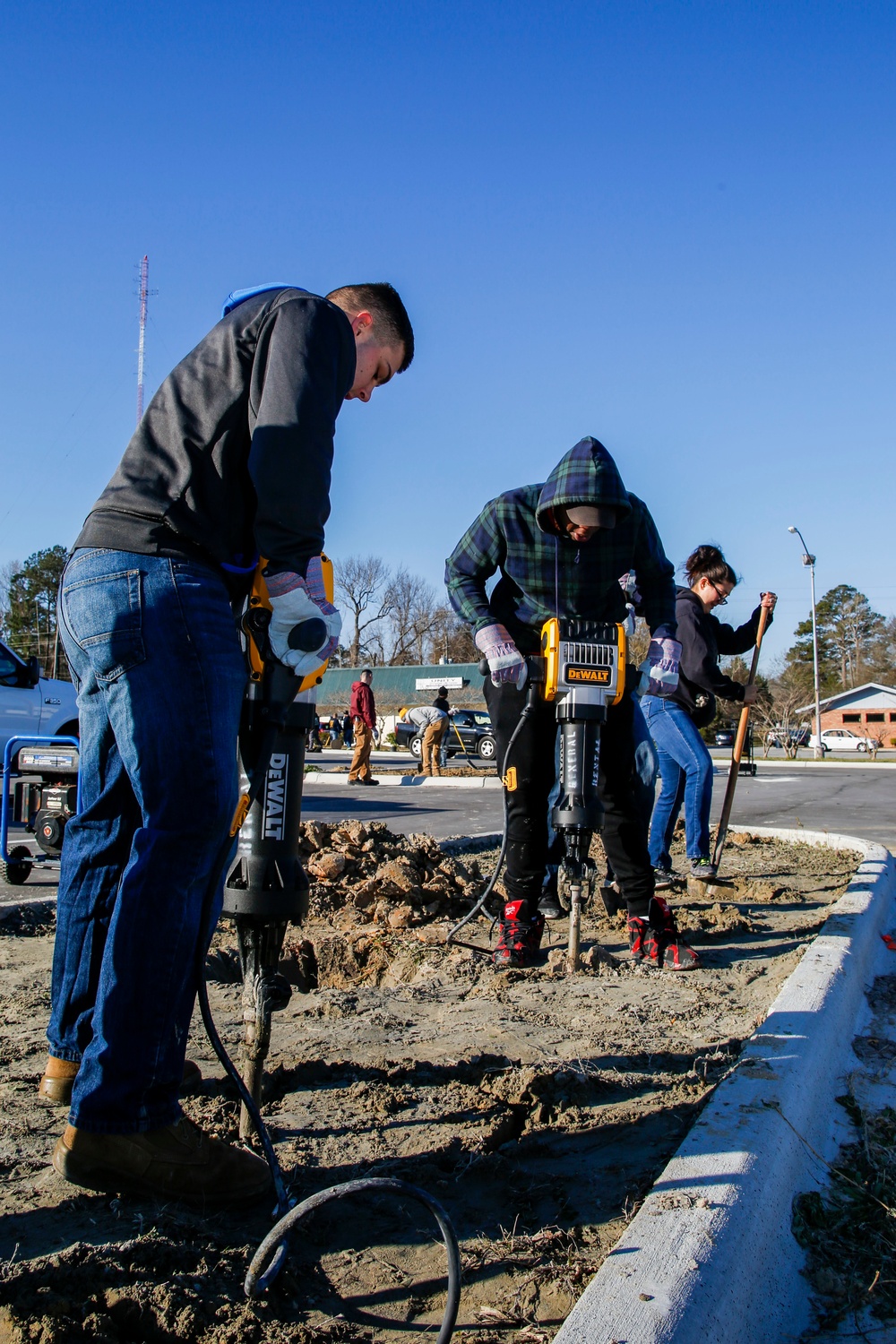 Three workers are engaged in ground-breaking or demolition work in what appears to be a parking lot or landscaped median area. Two individuals are operating DeWalt electric jackhammers/demolition hammers to break up compacted soil or concrete, while a third person assists with a shovel. Additional workers are visible in the background performing similar tasks.