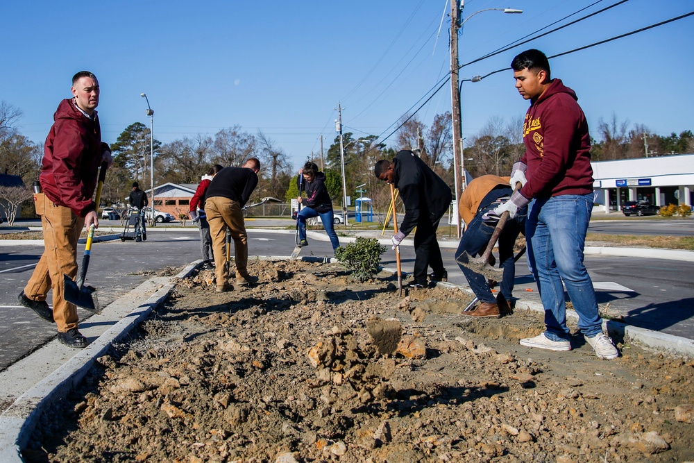 A group of people are engaged in outdoor landscaping or community improvement work in a parking lot median or island area. They are digging and moving soil using shovels and other hand tools, with at least one small shrub or plant visible in the disturbed earth. The setting appears to be a suburban commercial area on a clear, cool day.