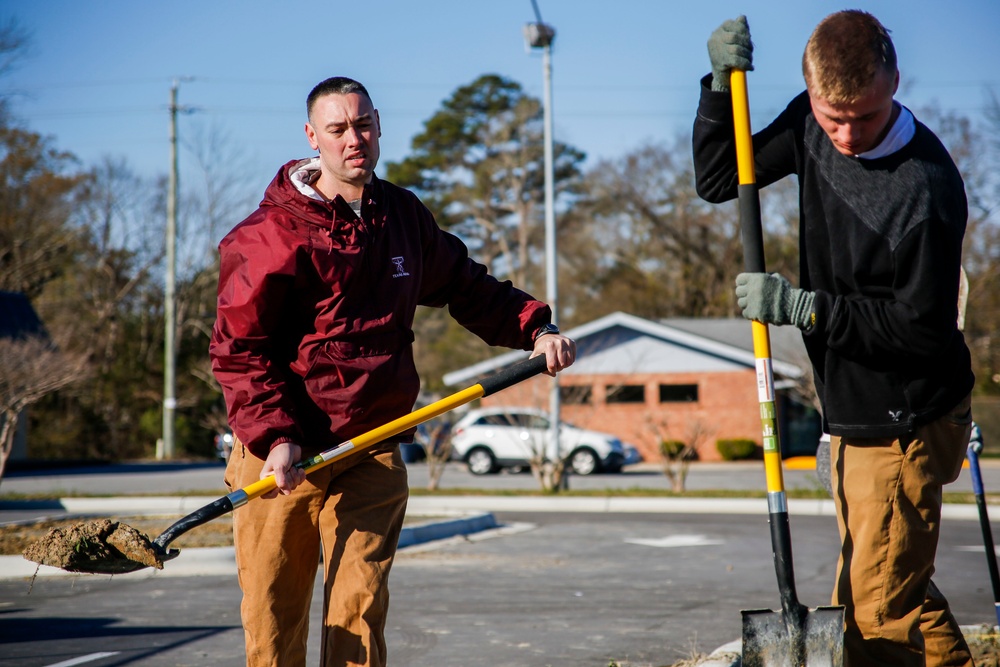 Two young men are performing outdoor manual labor using shovels with yellow and black handles in what appears to be a parking lot area. The man on the left wears a maroon jacket and tan work pants, while the man on the right wears a black sweatshirt with work gloves and matching tan pants. The setting appears to be a suburban area on a clear, sunny day with bare winter trees visible in the background.