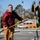 Arbor Day "Adopt an Island" Two young men are performing outdoor manual labor using shovels with yellow and black handles in what appears to be a parking lot area. The man on the left wears a maroon jacket and tan work pants, while the man on the right wears a black sweatshirt with work gloves and matching tan pants. The setting appears to be a suburban area on a clear, sunny day with bare winter trees visible in the background.