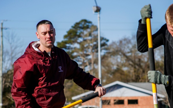 Two young men are performing outdoor manual labor using shovels with yellow and black handles in what appears to be a parking lot area. The man on the left wears a maroon jacket and tan work pants, while the man on the right wears a black sweatshirt with work gloves and matching tan pants. The setting appears to be a suburban area on a clear, sunny day with bare winter trees visible in the background.
