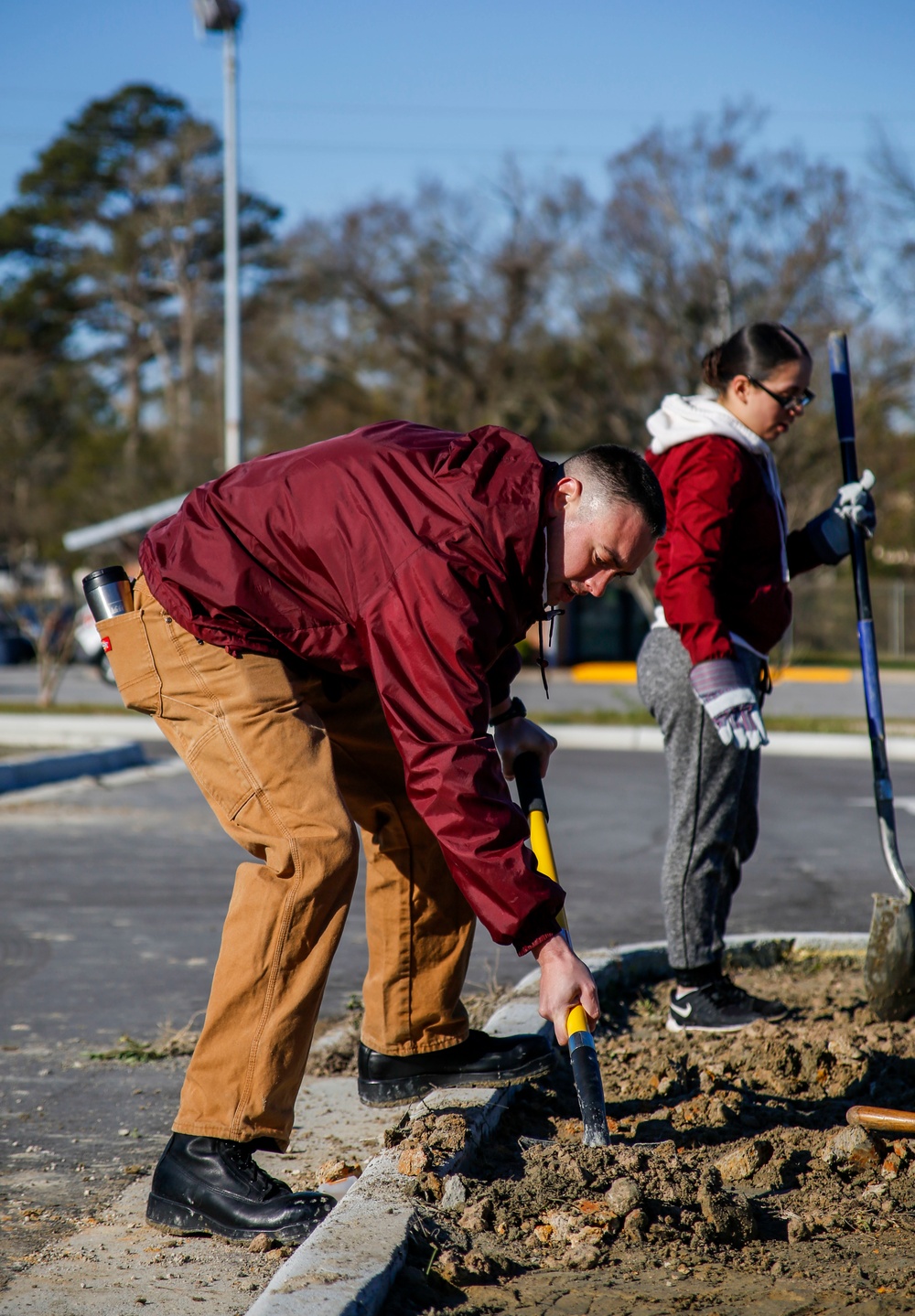 Two individuals are performing outdoor manual labor, likely landscaping or ground maintenance work near a parking lot or roadway. The person in the foreground, wearing a maroon jacket and tan work pants, is actively digging with a yellow-handled shovel into rocky, disturbed soil. A second person in the background, wearing a red top and gray pants with work gloves, holds a long-handled tool such as a rake or broom.
