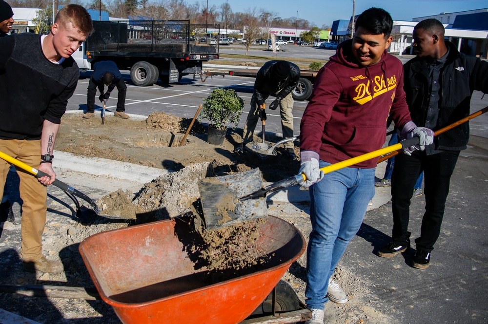 A group of young men are engaged in outdoor landscaping or construction work in a parking lot area. They are shoveling dirt and debris into a red wheelbarrow, with additional workers visible in the background digging around a small shrub or plant. The setting appears to be a commercial area with a flatbed truck and retail stores visible in the background.
