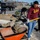 A group of young men are engaged in outdoor landscaping or construction work in a parking lot area. They are shoveling dirt and debris into a red wheelbarrow, with additional workers visible in the background digging around a small shrub or plant. The setting appears to be a commercial area with a flatbed truck and retail stores visible in the background.