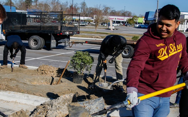 A group of young men are engaged in outdoor landscaping or construction work in a parking lot area. They are shoveling dirt and debris into a red wheelbarrow, with additional workers visible in the background digging around a small shrub or plant. The setting appears to be a commercial area with a flatbed truck and retail stores visible in the background.