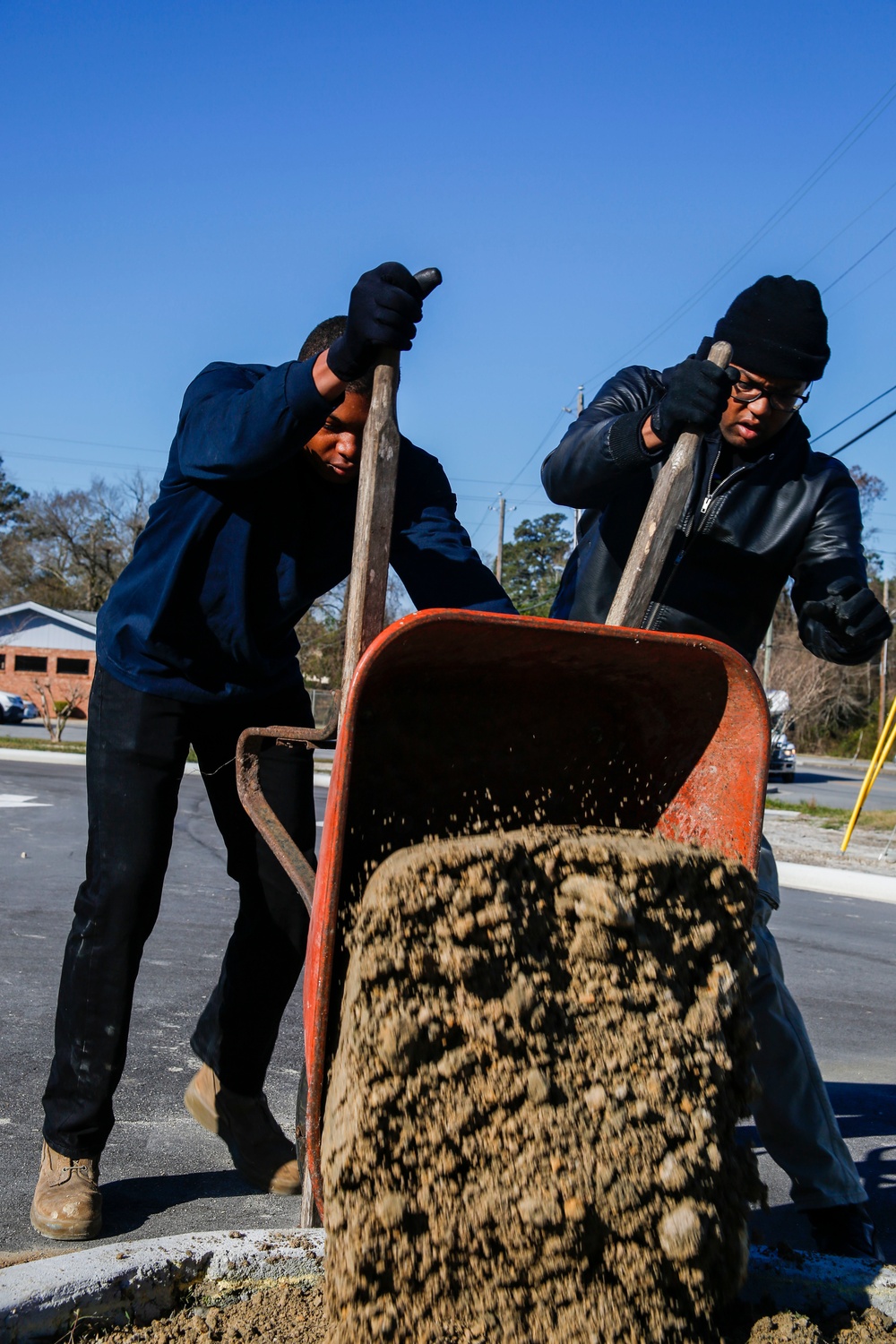 Two workers dressed in dark clothing and black gloves are actively dumping a load of gravel or sandy material from a red wheelbarrow onto a paved surface. The action is captured mid-motion, with material visibly cascading out of the tilted wheelbarrow. The setting appears to be an outdoor work site on a clear, sunny day with a residential area visible in the background.