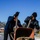 Two workers dressed in dark clothing and black gloves are actively dumping a load of gravel or sandy material from a red wheelbarrow onto a paved surface. The action is captured mid-motion, with material visibly cascading out of the tilted wheelbarrow. The setting appears to be an outdoor work site on a clear, sunny day with a residential area visible in the background.