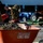 Two workers are engaged in outdoor landscaping or construction work, with one shoveling soil or dirt into a red wheelbarrow in the foreground. In the background, multiple tractors — including a red and a blue one — are parked in what appears to be a paved lot near a building. The scene suggests an active work site involving earth-moving or planting activities.