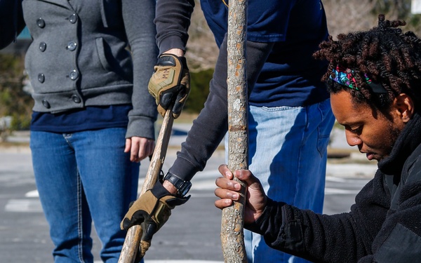 A group of people are planting a young tree outdoors in what appears to be a community or volunteer tree-planting effort. One person crouches while steadying the tree trunk with both hands, while another uses a shovel to move soil into the hole. A third person stands nearby observing or assisting.