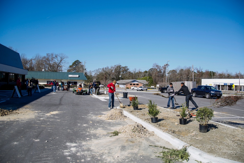A group of people are engaged in landscaping work in a commercial parking lot area on a clear, sunny day. Workers are using shovels to dig holes and plant potted shrubs along a newly constructed concrete median or curbed island. The scene appears to be a community or commercial property improvement project.