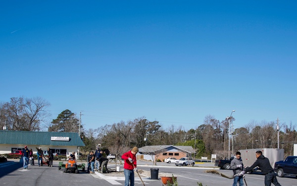 A group of people are engaged in landscaping work in a commercial parking lot area on a clear, sunny day. Workers are using shovels to dig holes and plant potted shrubs along a newly constructed concrete median or curbed island. The scene appears to be a community or commercial property improvement project.