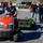 A group of workers wearing matching navy blue 'FMTB-E STAFF' shirts are engaged in landscaping or grounds maintenance work in a parking lot area. A young man in a camouflage cap is driving a red Husqvarna riding lawn tractor pulling a green utility trailer loaded with soil or mulch. Two other staff members are walking behind the trailer, and a fourth person in pink is visible in the background.