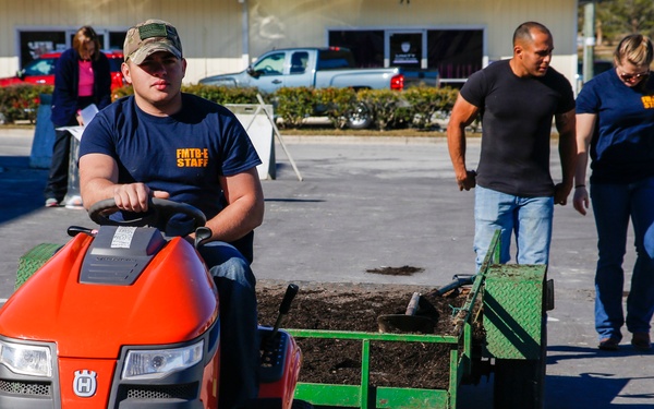 A group of workers wearing matching navy blue 'FMTB-E STAFF' shirts are engaged in landscaping or grounds maintenance work in a parking lot area. A young man in a camouflage cap is driving a red Husqvarna riding lawn tractor pulling a green utility trailer loaded with soil or mulch. Two other staff members are walking behind the trailer, and a fourth person in pink is visible in the background.