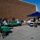 An outdoor community event or food distribution setup is taking place in a parking lot beside a large brick building. Multiple volunteers are working at tables covered with green tablecloths, with coolers, bins, and boxes of supplies visible. A blue pop-up canopy tent provides shade over one section of the operation.