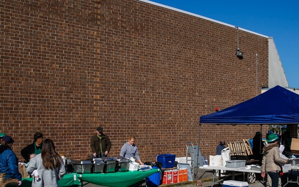 An outdoor community event or food distribution setup is taking place in a parking lot beside a large brick building. Multiple volunteers are working at tables covered with green tablecloths, with coolers, bins, and boxes of supplies visible. A blue pop-up canopy tent provides shade over one section of the operation.