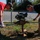 Two men are operating a gas-powered post hole digger (auger) on a grassy area near a street intersection. One man wears a red shirt and work gloves, while the other wears a black shirt and khaki pants. They appear to be drilling into the ground near an existing metal pole, likely for installation or maintenance purposes.