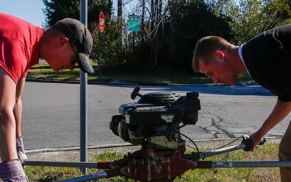 Two men are operating a gas-powered post hole digger (auger) on a grassy area near a street intersection. One man wears a red shirt and work gloves, while the other wears a black shirt and khaki pants. They appear to be drilling into the ground near an existing metal pole, likely for installation or maintenance purposes.