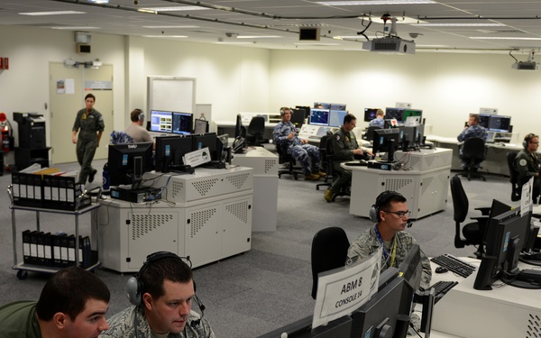 A military operations center or command and control facility showing personnel from multiple nations working at computer workstations. The room contains numerous consoles with monitors, and personnel are wearing a mix of US Air Force digital camouflage uniforms and what appear to be Australian Defence Force uniforms, suggesting a joint multinational exercise or operation. A console label reading 'ABM 8 CONSOLE 14' is visible in the foreground.