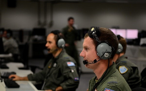 Military personnel in olive green flight suits are working at computer workstations in what appears to be an operations or command center. The individuals are wearing headsets with boom microphones, suggesting active communications. Shoulder patches visible on multiple personnel display the Australian flag, identifying them as Royal Australian Air Force (RAAF) members.