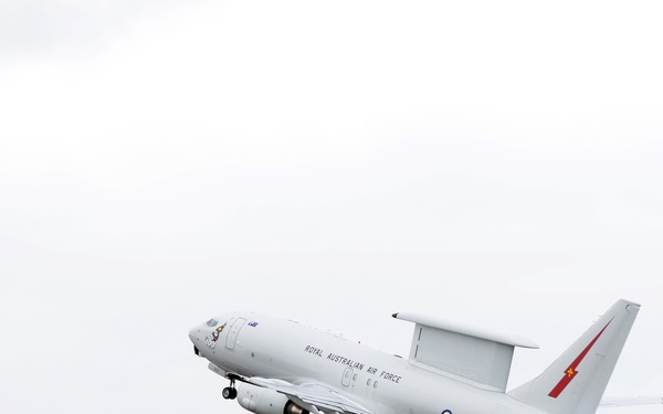 A Royal Australian Air Force (RAAF) E-7A Wedgetail airborne early warning and control aircraft is captured during takeoff or shortly after liftoff. The white military aircraft features a distinctive rotodome radar system mounted above the fuselage and displays RAAF markings including a roundel and lightning bolt tail design. The image was taken at what appears to be an Australian military or civilian airfield with buildings and trees visible in the background.