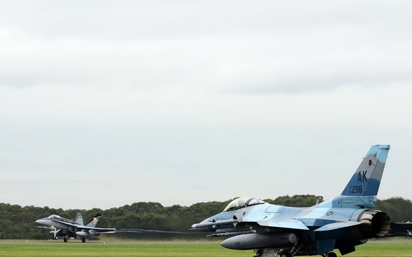 Two military fighter jets are taxiing on a runway at an airfield surrounded by green grass and trees under an overcast sky. The foreground aircraft is an F-16 Fighting Falcon bearing the tail code 'AK' and serial number '298', indicating it belongs to the Alaska Air National Guard or an Alaskan-based unit. A second aircraft, appearing to be an F/A-18 Hornet, is visible in the background also on the runway.