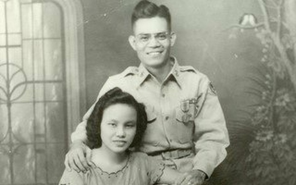 A formal studio portrait photograph, likely from the 1940s, showing a young man in military uniform seated with a young woman in front of him. The man has his arm around the woman in an affectionate pose, suggesting they are a couple. The sepia-toned image features a painted studio backdrop typical of mid-20th century portrait photography.