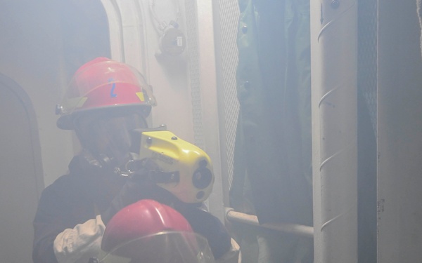 Two firefighters in full protective gear are navigating through a smoke-filled corridor, likely during a firefighting drill or active emergency response. The lead firefighter wears a red helmet marked with the number '2' and carries a yellow thermal imaging camera. The environment appears to be a confined industrial or naval vessel passageway filled with dense smoke.