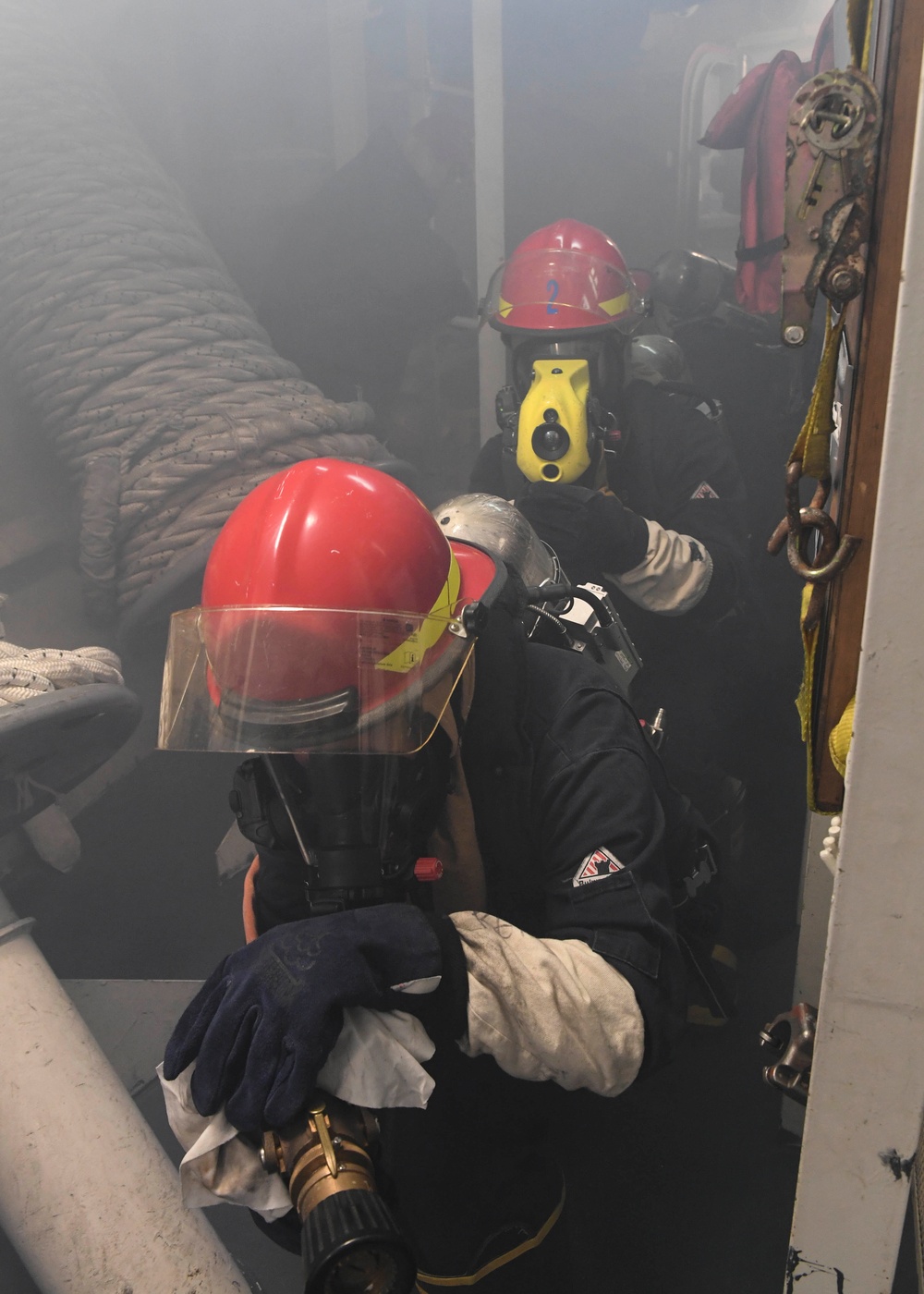 Two firefighters or emergency response personnel in full protective gear, including red helmets, face shields, breathing apparatus (SCBA), and fire-resistant suits, are operating in a smoke-filled environment aboard what appears to be a vessel or ship. The foreground figure is crouching and holding a brass fire hose nozzle, while a second figure marked with the number '2' on their helmet is visible behind. The scene strongly suggests a maritime firefighting drill or active fire suppression operation.
