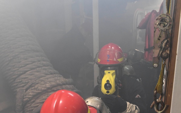Two firefighters or emergency response personnel in full protective gear, including red helmets, face shields, breathing apparatus (SCBA), and fire-resistant suits, are operating in a smoke-filled environment aboard what appears to be a vessel or ship. The foreground figure is crouching and holding a brass fire hose nozzle, while a second figure marked with the number '2' on their helmet is visible behind. The scene strongly suggests a maritime firefighting drill or active fire suppression operation.
