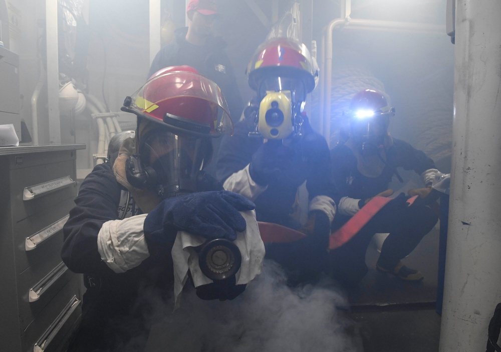 The image shows a group of personnel in firefighting or damage control gear operating in a smoke-filled interior space, likely aboard a naval vessel or industrial facility. They are wearing red helmets with face shields, protective gloves, and appear to be conducting a firefighting drill or responding to an emergency. One individual has a mounted flashlight illuminating the smoky environment.