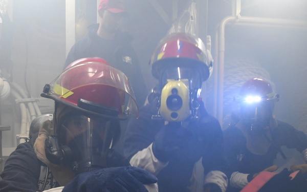 The image shows a group of personnel in firefighting or damage control gear operating in a smoke-filled interior space, likely aboard a naval vessel or industrial facility. They are wearing red helmets with face shields, protective gloves, and appear to be conducting a firefighting drill or responding to an emergency. One individual has a mounted flashlight illuminating the smoky environment.