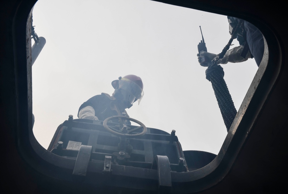 The image is taken from a low-angle perspective looking upward through a hatch or opening, showing two individuals in protective gear. One person wearing a red helmet and face shield appears to be operating controls with a steering wheel visible, while a second person holds a handheld radio/walkie-talkie. The scene appears to involve emergency response or industrial operations, with hazy or smoky atmospheric conditions visible.