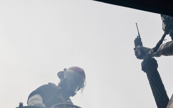 The image is taken from a low-angle perspective looking upward through a hatch or opening, showing two individuals in protective gear. One person wearing a red helmet and face shield appears to be operating controls with a steering wheel visible, while a second person holds a handheld radio/walkie-talkie. The scene appears to involve emergency response or industrial operations, with hazy or smoky atmospheric conditions visible.