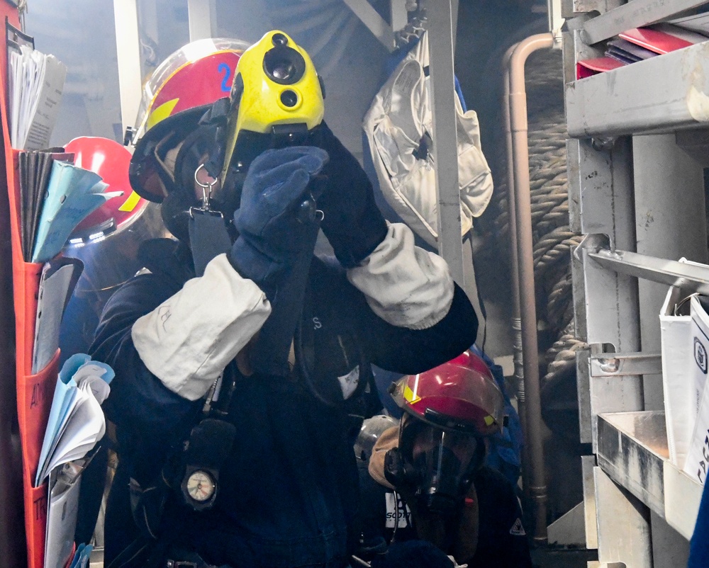 Two firefighters or damage control personnel are navigating a smoke-filled interior space, likely aboard a naval vessel or industrial facility. The lead figure wears a yellow-fronted red helmet marked with the number '2' and is adjusting a breathing apparatus mask. The environment appears hazardous, with visible smoke and confined quarters filled with equipment and documents.