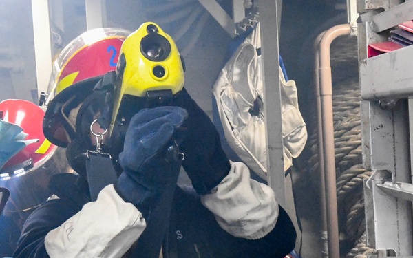 Two firefighters or damage control personnel are navigating a smoke-filled interior space, likely aboard a naval vessel or industrial facility. The lead figure wears a yellow-fronted red helmet marked with the number '2' and is adjusting a breathing apparatus mask. The environment appears hazardous, with visible smoke and confined quarters filled with equipment and documents.