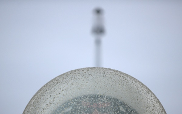 A close-up view of a rain-covered military jet fighter canopy, shot from the nose looking rearward. Through the wet, moisture-beaded canopy, a red star insignia is visible on what appears to be the pilot's helmet or cockpit interior, suggesting a Russian or Chinese military aircraft. A vertical antenna or mast is visible in the blurred background against an overcast grey sky.