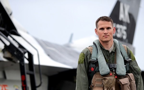 A military pilot in full flight gear stands on an airfield tarmac in front of a fighter jet. The pilot is wearing an olive green flight suit, survival vest, and life preserver unit, and is carrying a flight bag. The aircraft visible in the background displays tail markings 'AK' and the number '375', consistent with U.S. Air Force markings.