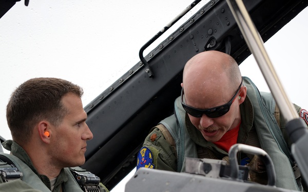 Two military aviators in olive drab flight suits are interacting in or around the cockpit of a military jet aircraft. One pilot is seated in the cockpit while the other leans in from outside, appearing to discuss cockpit instruments or controls. The open canopy is visible overhead, and both individuals wear standard flight gear including survival vests.
