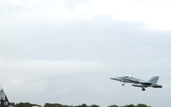 A military airfield scene showing two jet aircraft — one taxiing on a wet runway and one airborne. The ground aircraft is an F-16 Fighting Falcon marked with tail code 'AK' and number '375', indicating Alaska Air National Guard or USAF Alaskan Command. The airborne aircraft appears to be an F/A-18 Hornet or Super Hornet in the process of taking off or landing.