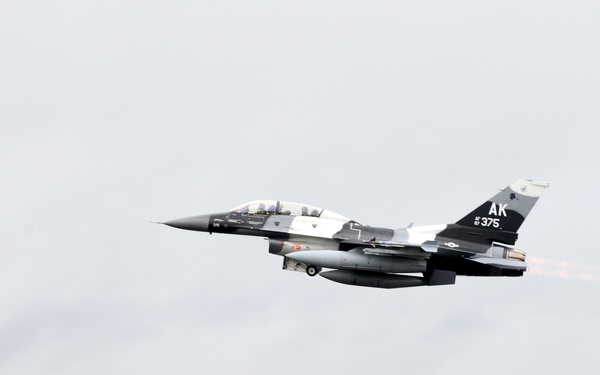 A U.S. Air Force F-16 Fighting Falcon jet, marked with tail code 'AK 375', is captured during takeoff or low-altitude flight just above a runway. The aircraft displays afterburner flame from its engine and carries underwing stores. The background shows a tree line and grassy airfield, with motion blur indicating high speed.