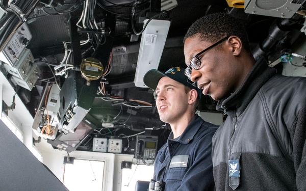 Two individuals are working together at a control console inside what appears to be a military vessel's bridge or command station. One person is wearing a dark uniform with a cap, while the other wears a black fleece jacket with glasses and a watch. They appear to be reviewing or operating equipment on a monitor or control panel.