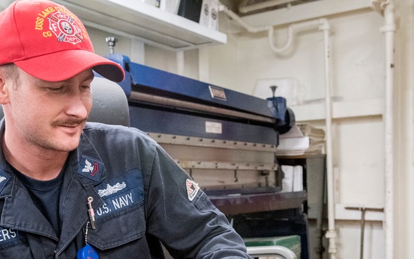 A U.S. Navy sailor named Bowers is seated aboard a naval vessel, wearing dark blue coveralls and a red baseball cap. He is holding what appear to be writing instruments or small tools in both hands and appears to be examining or working with them. The setting is an interior shipboard area with visible pipes, an exit door, and industrial equipment in the background.