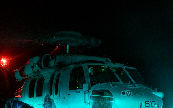 A military helicopter, marked with the number '613', is parked on what appears to be a naval vessel's flight deck at night. Ground crew personnel are working around the aircraft under teal/green artificial lighting. The scene captures nighttime flight deck operations with visible safety lighting and wet deck surfaces.