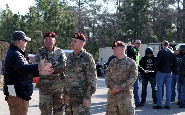 A civilian in casual attire appears to be briefing or conversing with three U.S. Army soldiers wearing OCP (Operational Camouflage Pattern) uniforms and maroon/crimson berets, indicative of Airborne units. The setting appears to be a parking area or training lot on a military installation, likely Fort Bragg (now Fort Liberty) based on the Airborne insignia. In the background, a group of people in motorcycle gear are gathered, suggesting a motorcycle safety or rider training event.