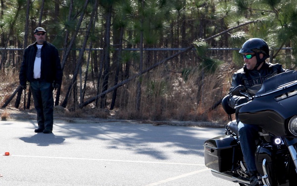 A rider on a black Harley-Davidson motorcycle navigates a marked course in a parking lot, with small red cones laid out in a pattern on the pavement. A second person stands nearby observing, likely serving as an instructor or evaluator. The setting appears to be a motorcycle safety or skills training course held outdoors near a wooded area.