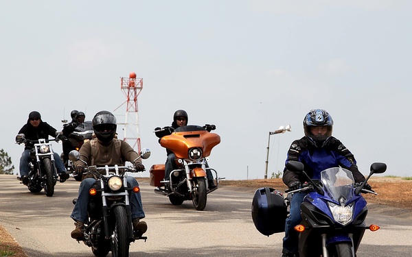A group of motorcyclists rides together in a convoy along a paved road, likely participating in an organized group ride or rally. The riders are traveling toward the camera on what appears to be a lightly trafficked road surrounded by sparse trees and dry grass. Various motorcycle styles are represented, ranging from sport bikes to cruisers and touring bikes.