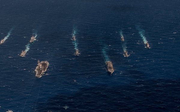 An aerial photograph captures a large naval formation consisting of multiple warships, including what appear to be at least two aircraft carriers, sailing in open ocean. Several military aircraft, including what appears to be a large bomber (likely a B-52) flanked by fighter jets, are flying in formation in the foreground. The ships are leaving visible wake trails, suggesting they are underway at speed in a coordinated exercise or demonstration.