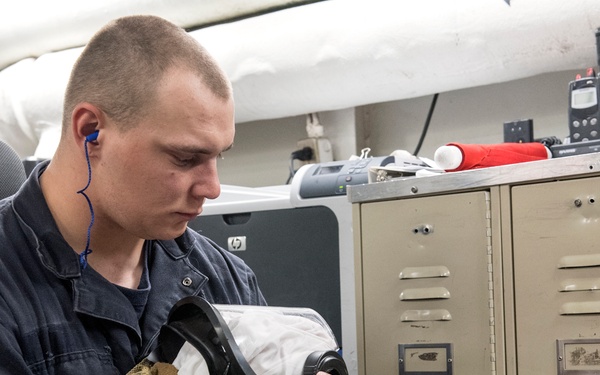 A young male service member in dark blue utility coveralls sits in an interior workspace, inspecting or maintaining what appears to be a gas mask or respirator with a tan mesh cover. The setting appears to be aboard a military vessel or installation, indicated by the metal lockers, overhead pipes, and military equipment visible in the background. He is wearing blue earbuds and is focused intently on the equipment in his hands.
