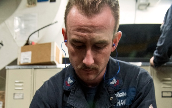 A U.S. Navy sailor named Bowers, wearing dark blue Navy coveralls, is kneeling on a red deck floor working on equipment. He appears to be handling a cylindrical canister or container with visible labeling. The setting appears to be an interior compartment of a naval vessel with metal lockers visible in the background.