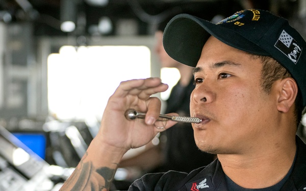 A U.S. Navy sailor named Pena is shown aboard what appears to be a naval vessel's bridge or combat information center, blowing a boatswain's pipe (bosun's whistle). The sailor is wearing standard U.S. Navy working uniform (NWU) with name tape, rating badges, and a ship's ball cap. The background shows navigation and control consoles typical of a naval ship's bridge.